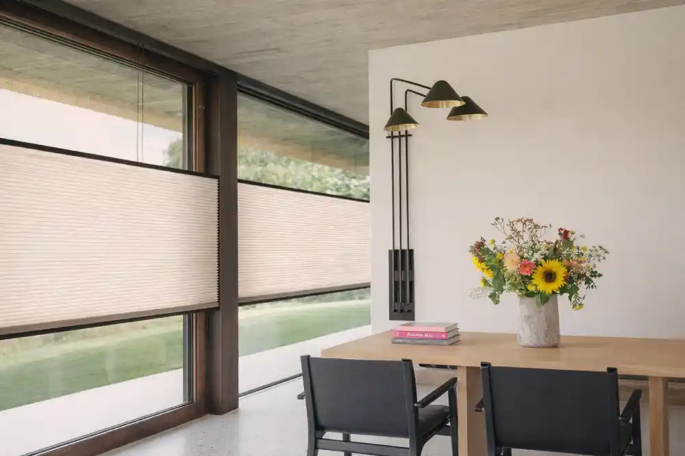 Bahçeye bakan geniş pencereli, gri plise perdeli ve kahverengi deri koltuklu modern oturma odası.